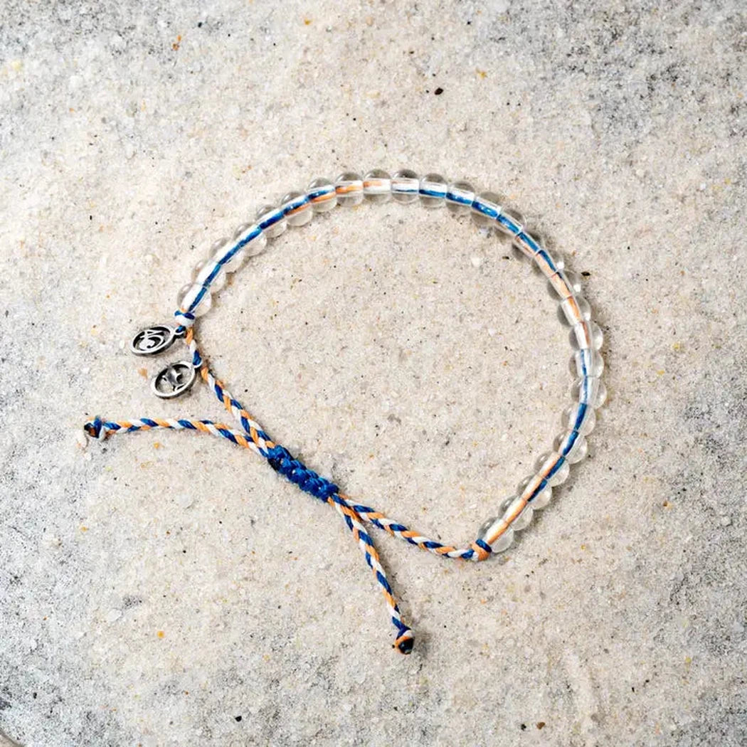 A close-up of the 4Ocean Reef Shark Beaded Bracelet - July 2024 Limited Edition on a light gray textured surface, showing the beaded design and blue and orange cord.