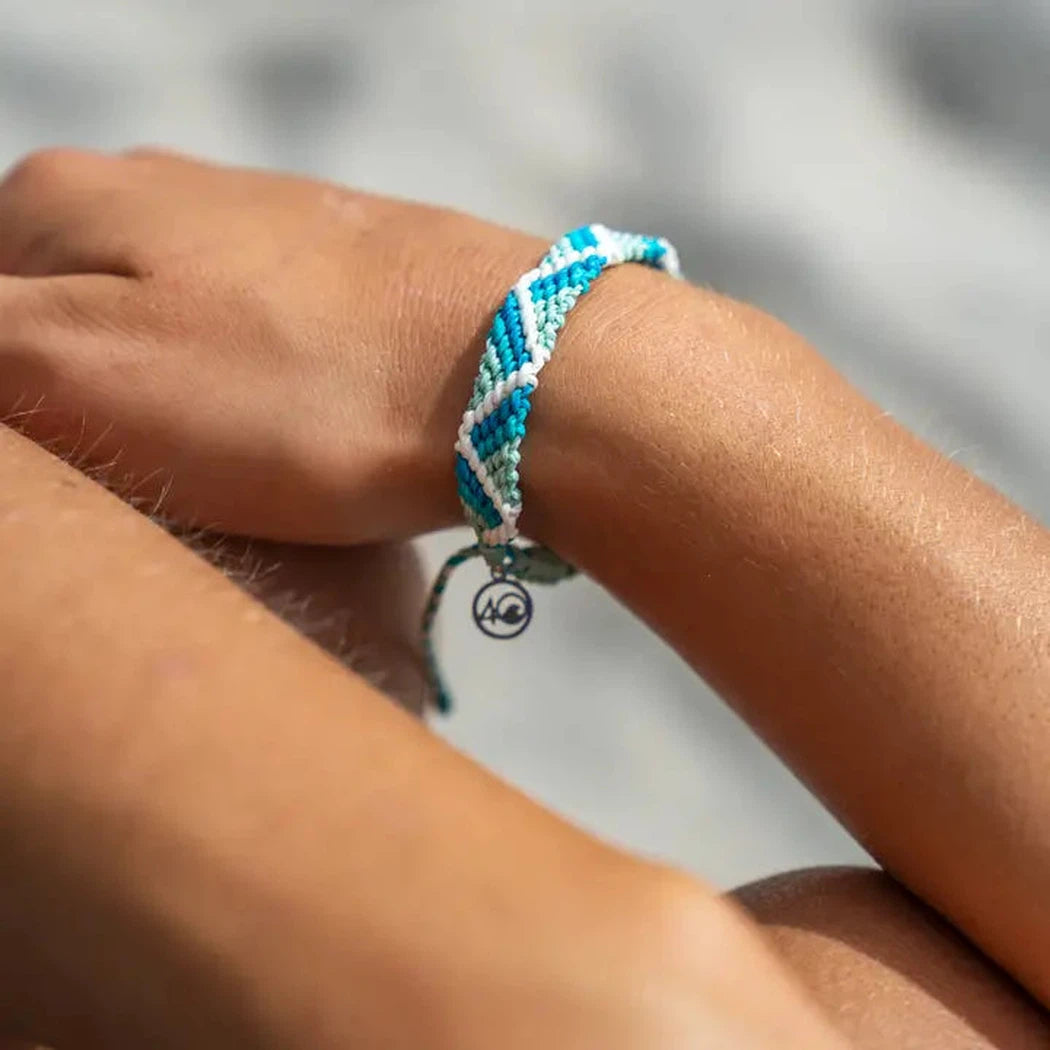 A close-up of a hand wearing the 4Ocean Riptide Bracelet in Mint Glow with a small charm, against a blurred outdoor background.