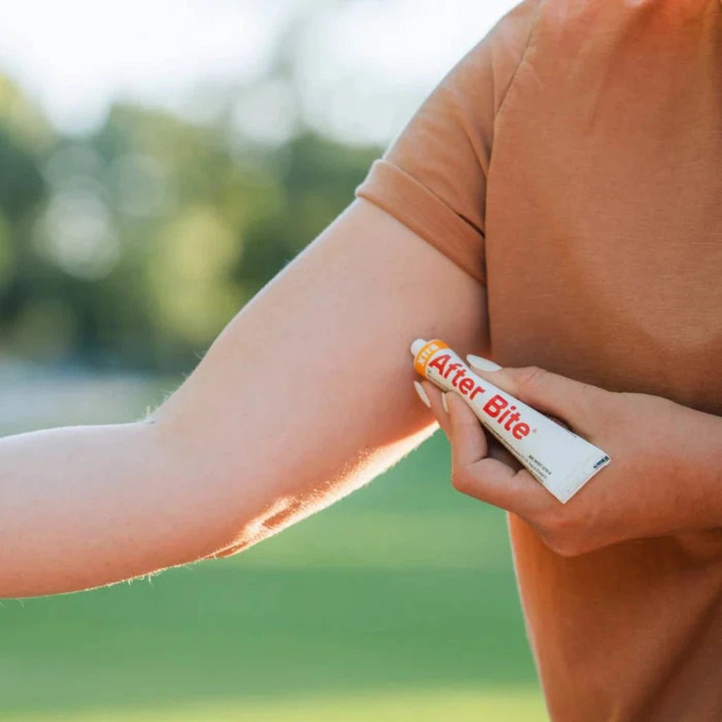 A person applies Adventure Medical Kits After Bite Xtra to their arm in an outdoor setting with green grass and trees.