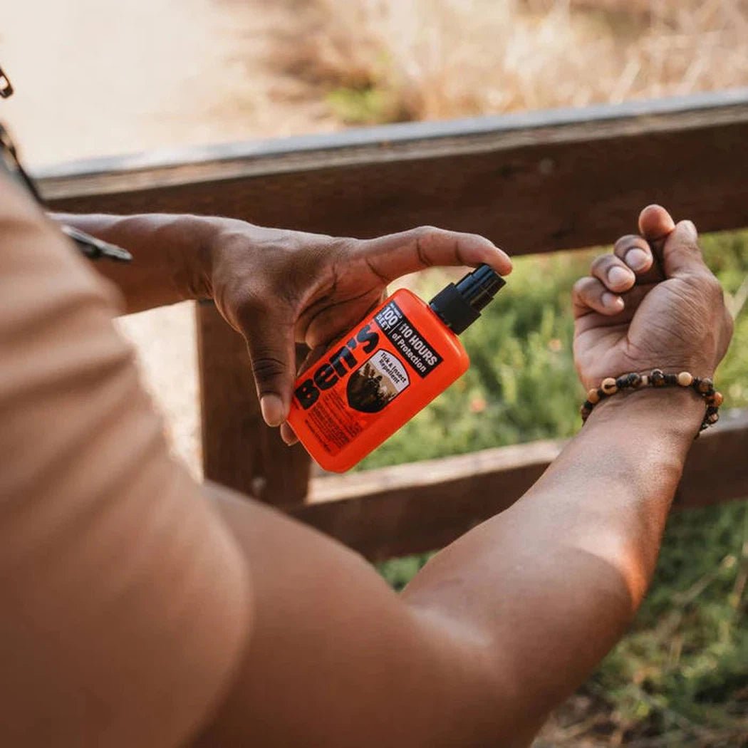 A person sprays Ben's 100 Tick and Insect Repellent on their arm while standing near a wooden fence with grass in the background.