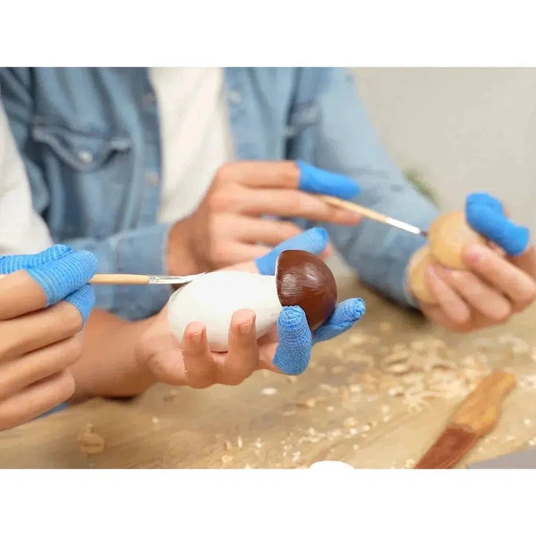 Two people wearing blue fingerless gloves use the Beavercraft Family Fun Wood Carving Kit to paint wooden shapes on a table.