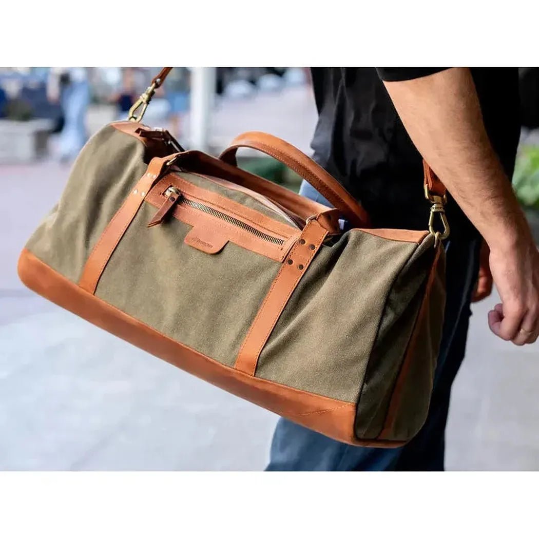 A man carries the Beavercraft Journey - Canvas and Leather Travel Luggage Duffel Bag in a side view with a city background.