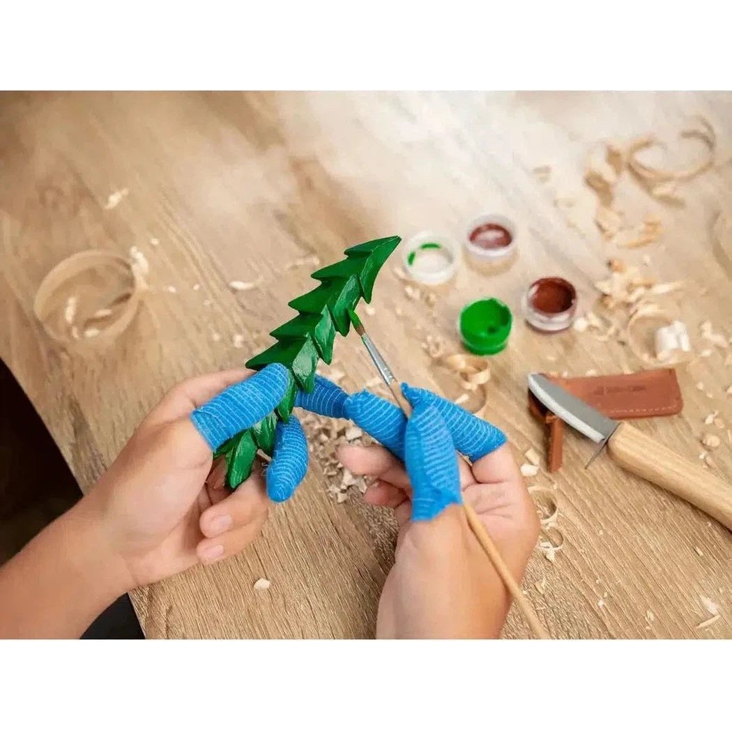 A person wearing blue gloves holds a green carved tree while painting it, with the Beavercraft Kids Carving Hobby Kit on a wood - filled workbench.