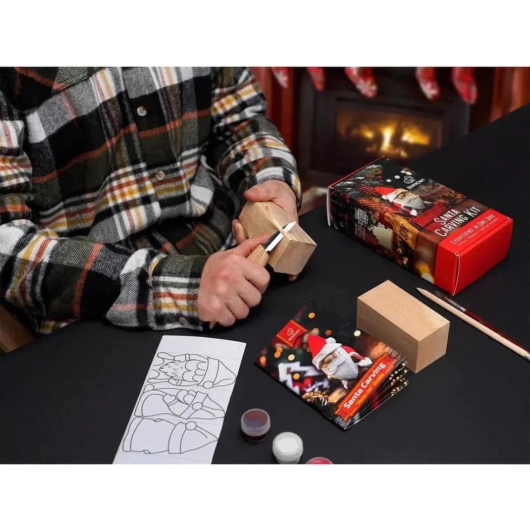 A person in a plaid shirt carves wood with the Beavercraft Santa Carving Hobby Kit on a black table near a fireplace.