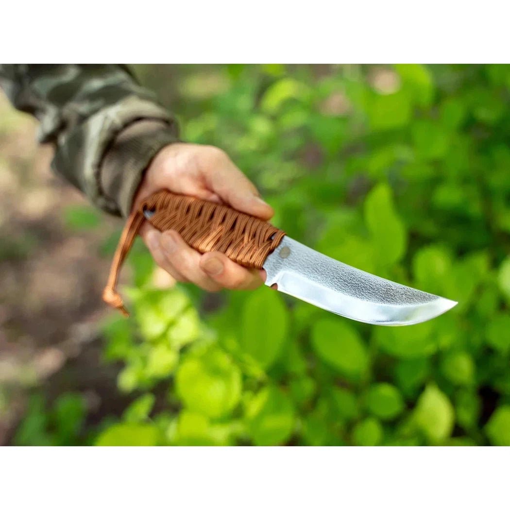 A hand holds the Beavercraft Skeleton Knife with a woven brown handle, showing the blade against a green leafy background.