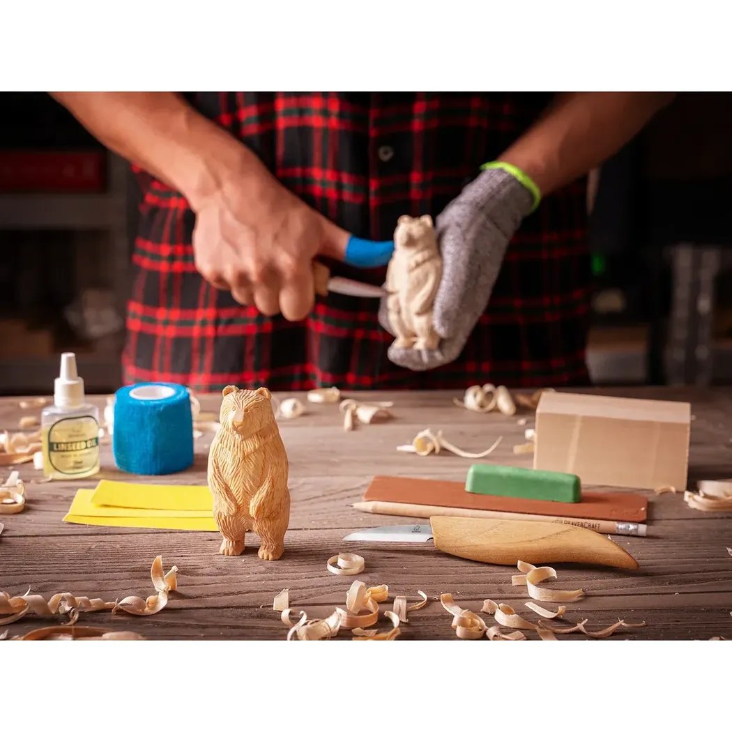 A person in a red plaid shirt carves a wooden bear with the Beavercraft Standing Bear Wood Carving kit on a cluttered workbench.