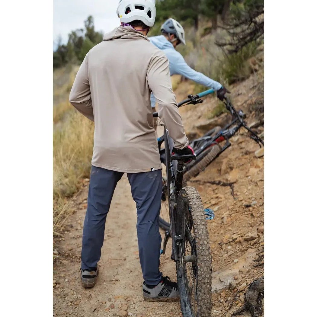 A man in a Dust Belong Designs Men's Core Traverse Tek Hoodie stands by a mountain bike on a dirt trail.
