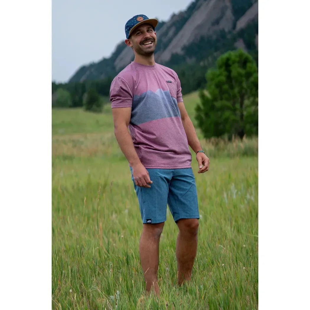 A smiling man in a Grandview Belong Designs Men's Graphic Traverse Tek Tee stands in a grassy field with mountains in the background.