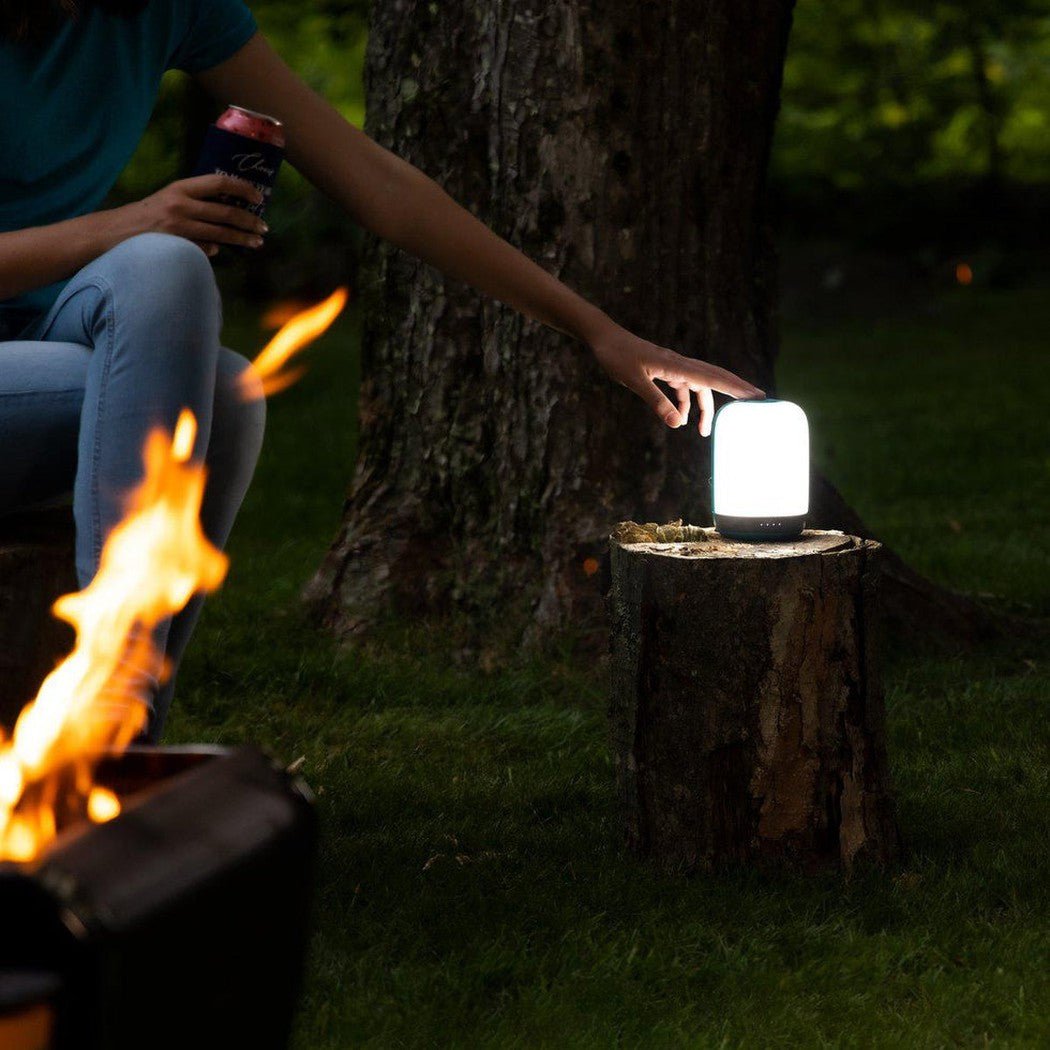 A person touches the BioLite Alpenglow 500 Lantern on a tree stump near a campfire at night.