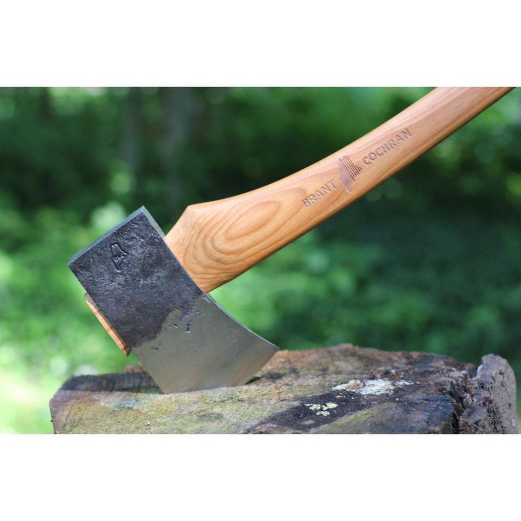 A close-up of the Brant & Cochran Allagash Cruiser Maine Wedge Camp Axe with a wooden handle and charcoal-colored blade resting on a tree stump in a green outdoor setting.