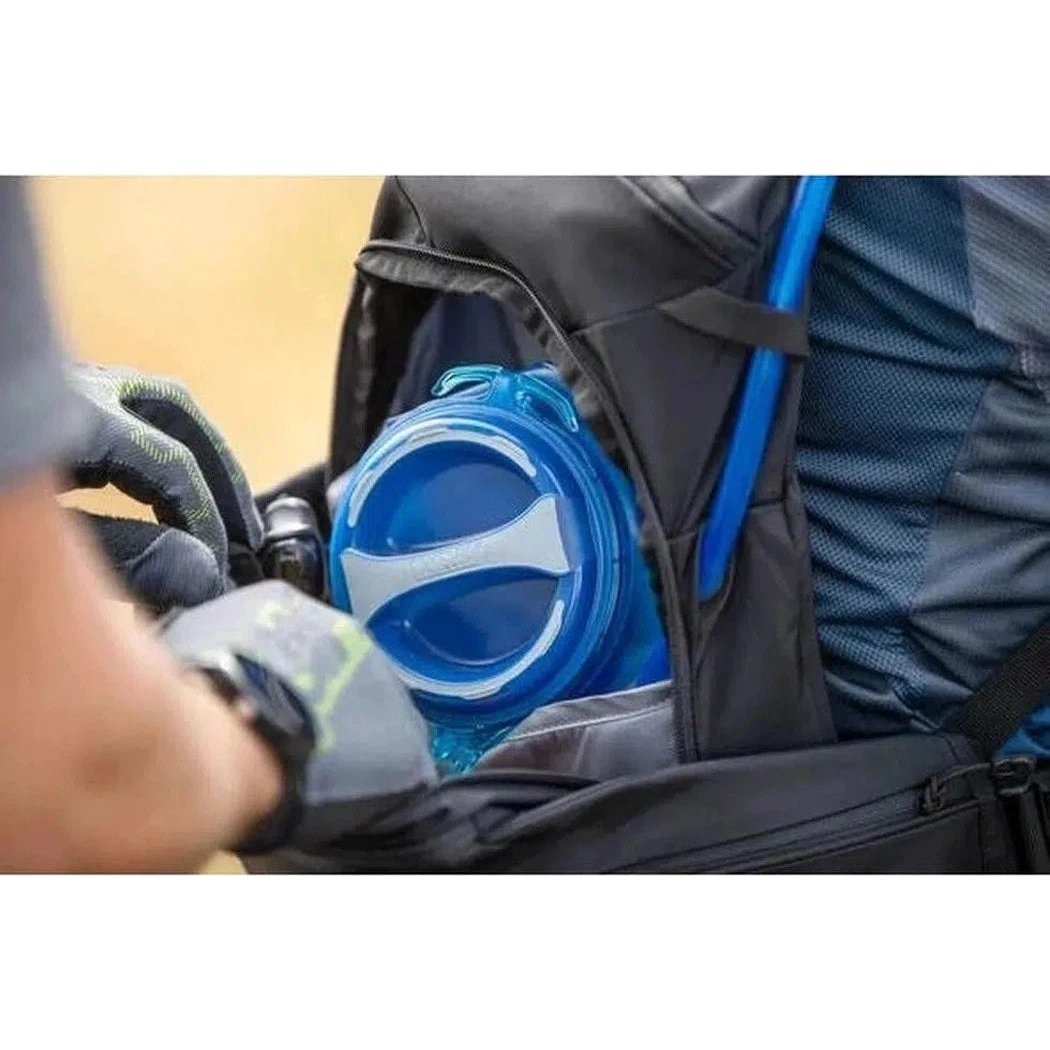 A close-up of the CamelBak Crux Reservoir On/Off Valve in a black backpack with a blue tube, showing the valve and a gloved hand adjusting it.