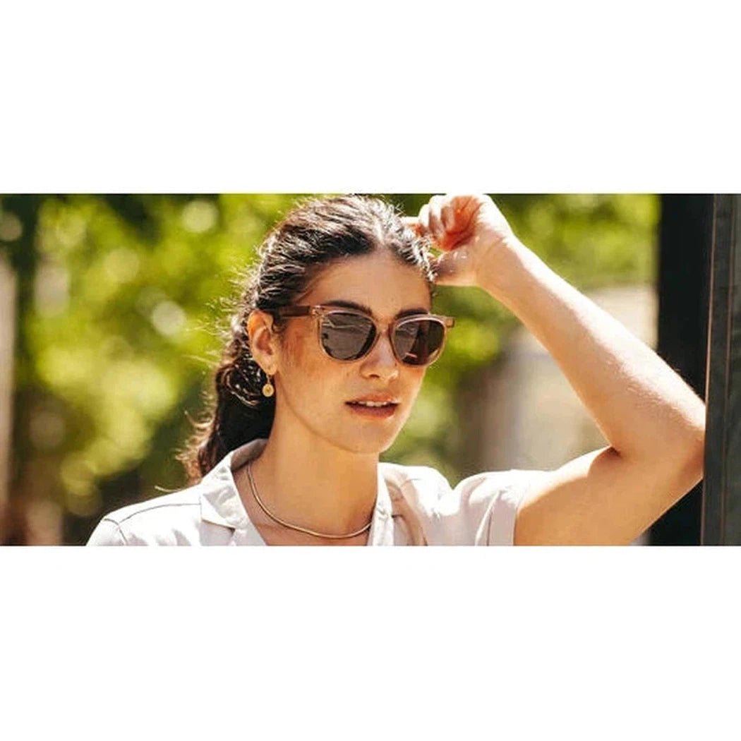A woman in a white shirt wears the Camp Eyewear Cove sunglasses with a green outdoor background.