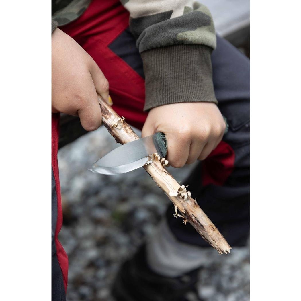 A person uses the Casstrom No 8 Swedish Forest Knife Green Micarta to carve a stick, with a rocky background visible.