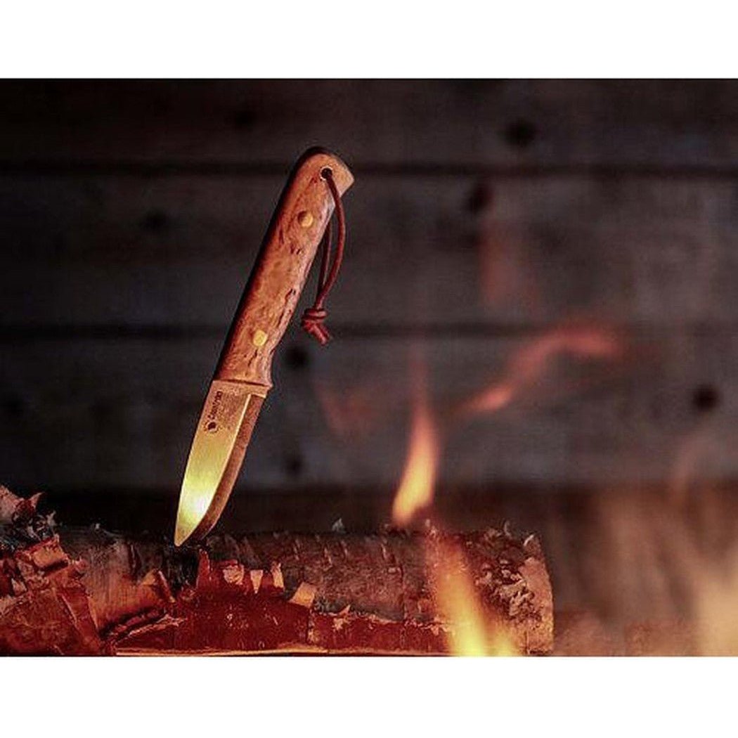 A Casstrom Woodsman knife with a Curly Birch handle sits on a burning log in front of a dark wooden wall.