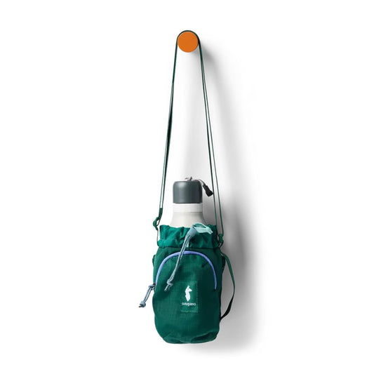 A green Cotopaxi Todo Water Bottle Sling hangs from an orange hook on a white wall.