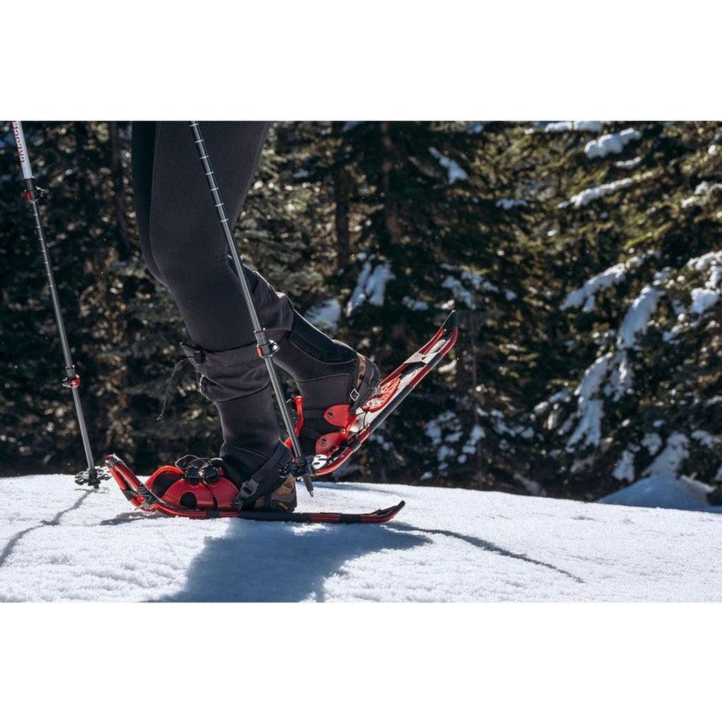 A person wearing black pants and boots stands on red Crescent Moon Yellowstone 24.5 snowshoes in a snowy forest.