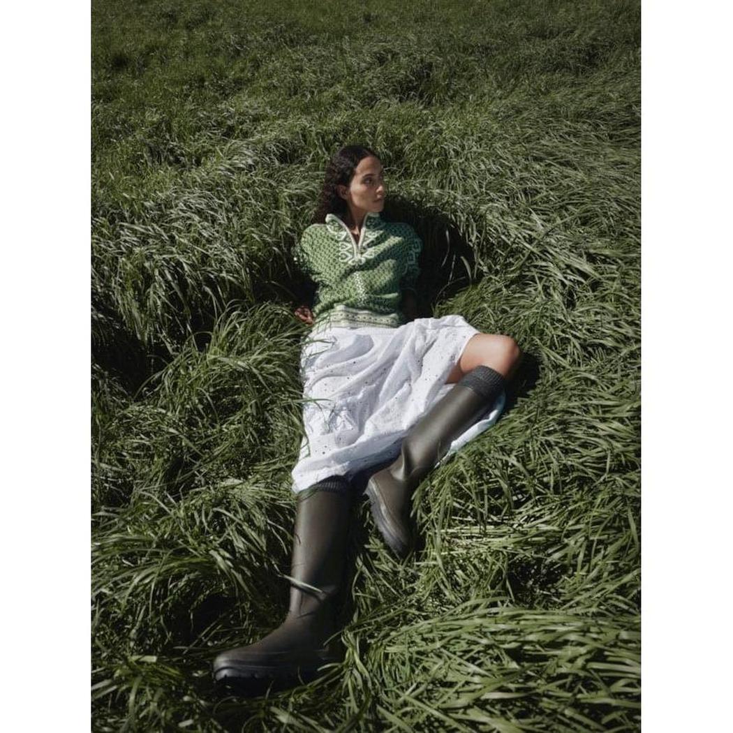 A woman wearing the Dale Of Norway Women's Leknes Sweater lies in tall green grass, showcasing the sweater and its offwhite skirt.