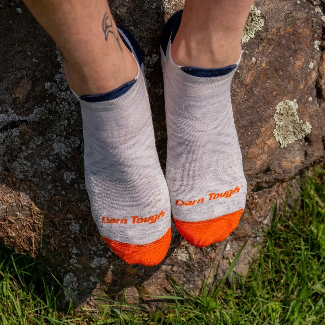 A close-up of a person wearing Darn Tough men's ash no show tab running socks with orange toes, sitting on a rock with grass below.