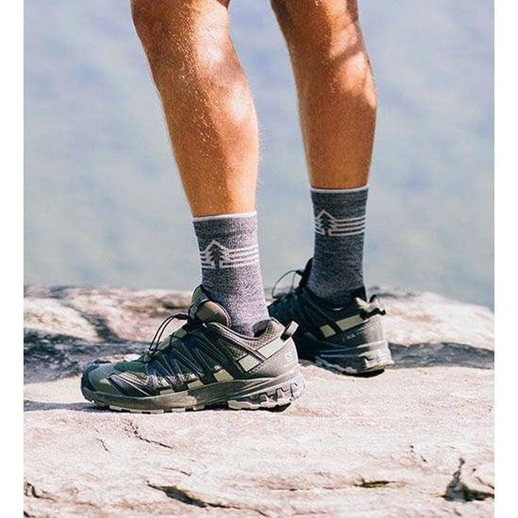 A person wearing FITS Light Hiker (Gifford) Crew socks and shoes stands on a rocky surface with a mountain background.