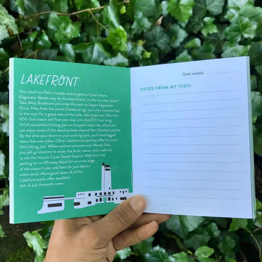 A hand holds the Free Period Press Cleveland Metroparks Mini-Guide open to a green "Lakefront" page with a white "Notes from my visit" page, against a background of green leaves.