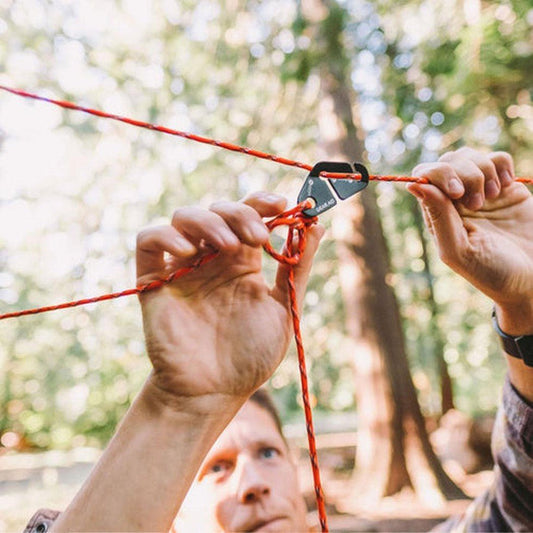 A person ties a red rope with a black carabiner from the Gear Aid Camp Line Kit in a forest setting.