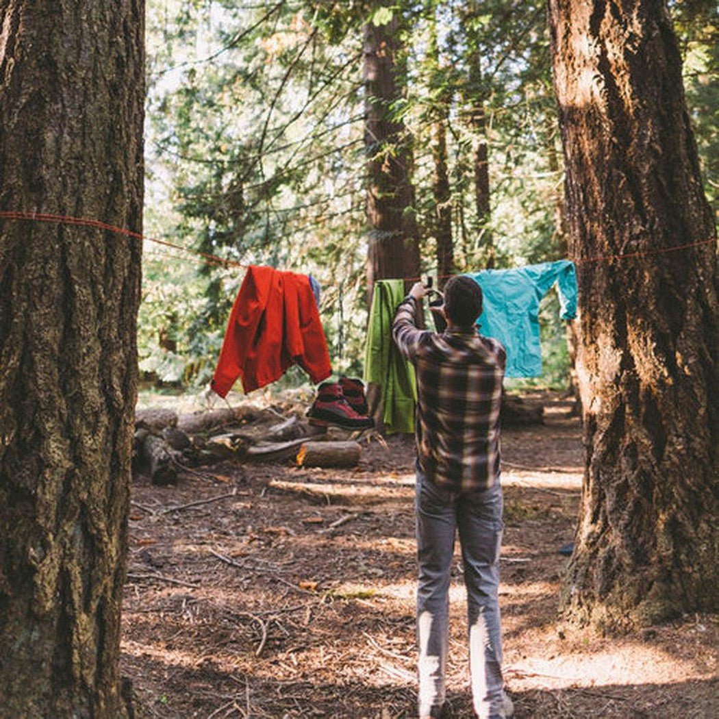 A man hangs colorful clothes on a line between trees while wearing the Gear Aid Camp Line Kit in a forest setting.