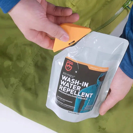 A person holds a 10oz Gear Aid Revivex Wash-In Water Repellent pouch with an orange cap, against a green fabric background.