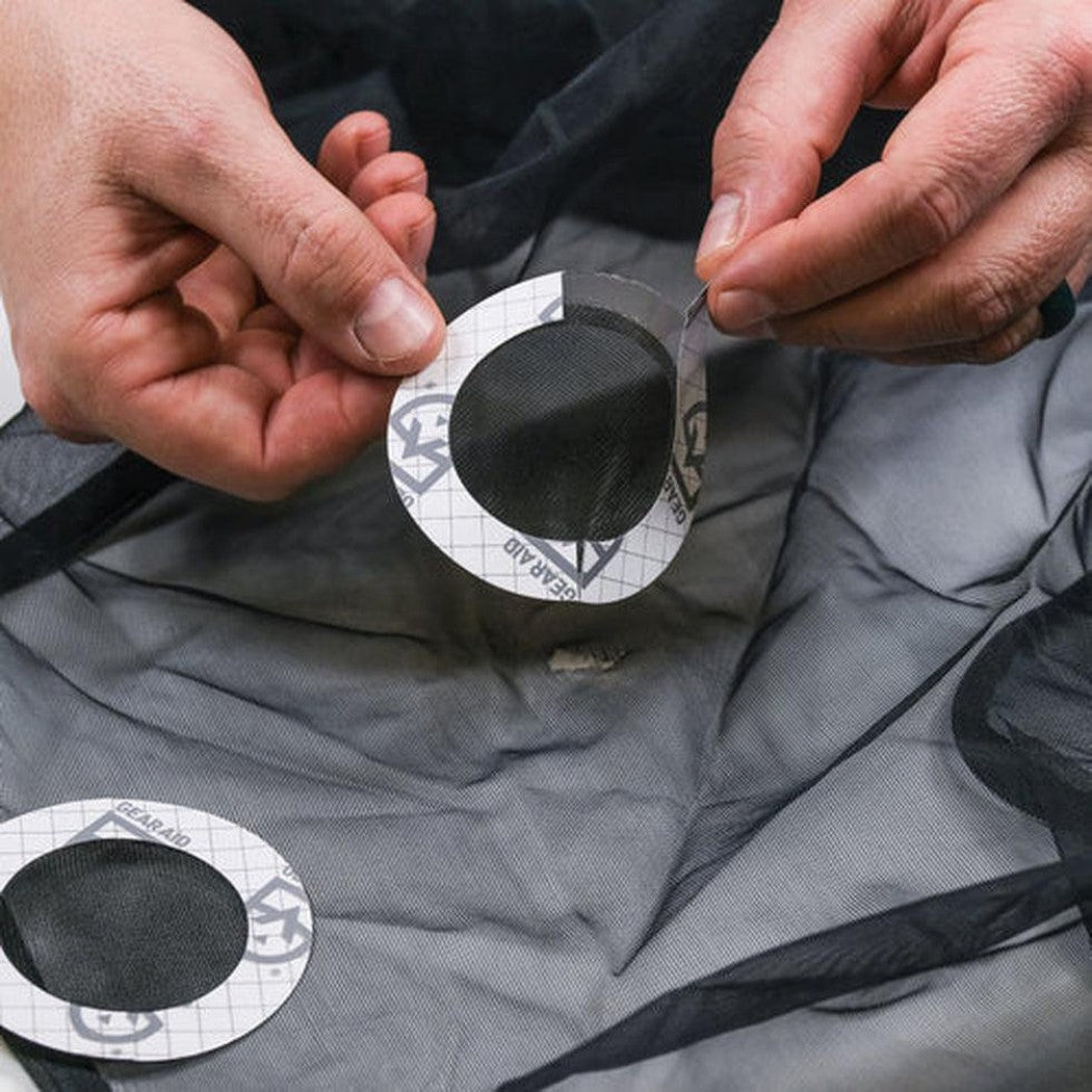 Two hands hold a Gear Aid Tenacious Tape Mesh Patch over a charcoal mesh fabric, with another patch visible below.
