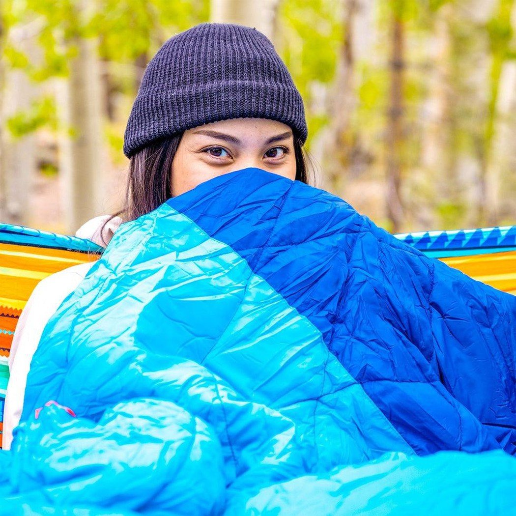 A woman wearing a charcoal beanie holds a Grand Trunk 360 ThermaQuilt in front of her face, with a forest background.