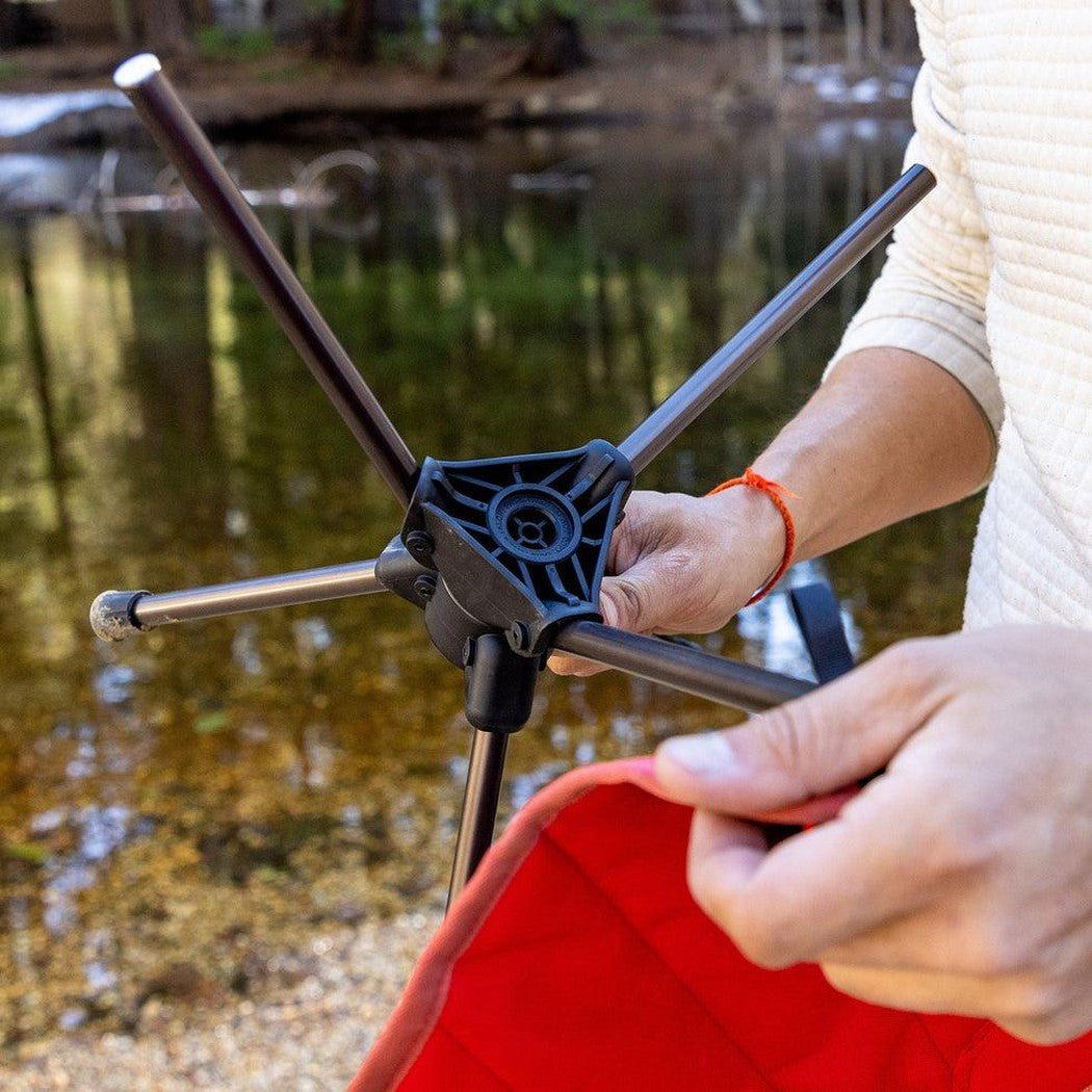 A person holds the Grand Trunk Compass 360 Stool, showing its black frame and red seat, against a lake background.