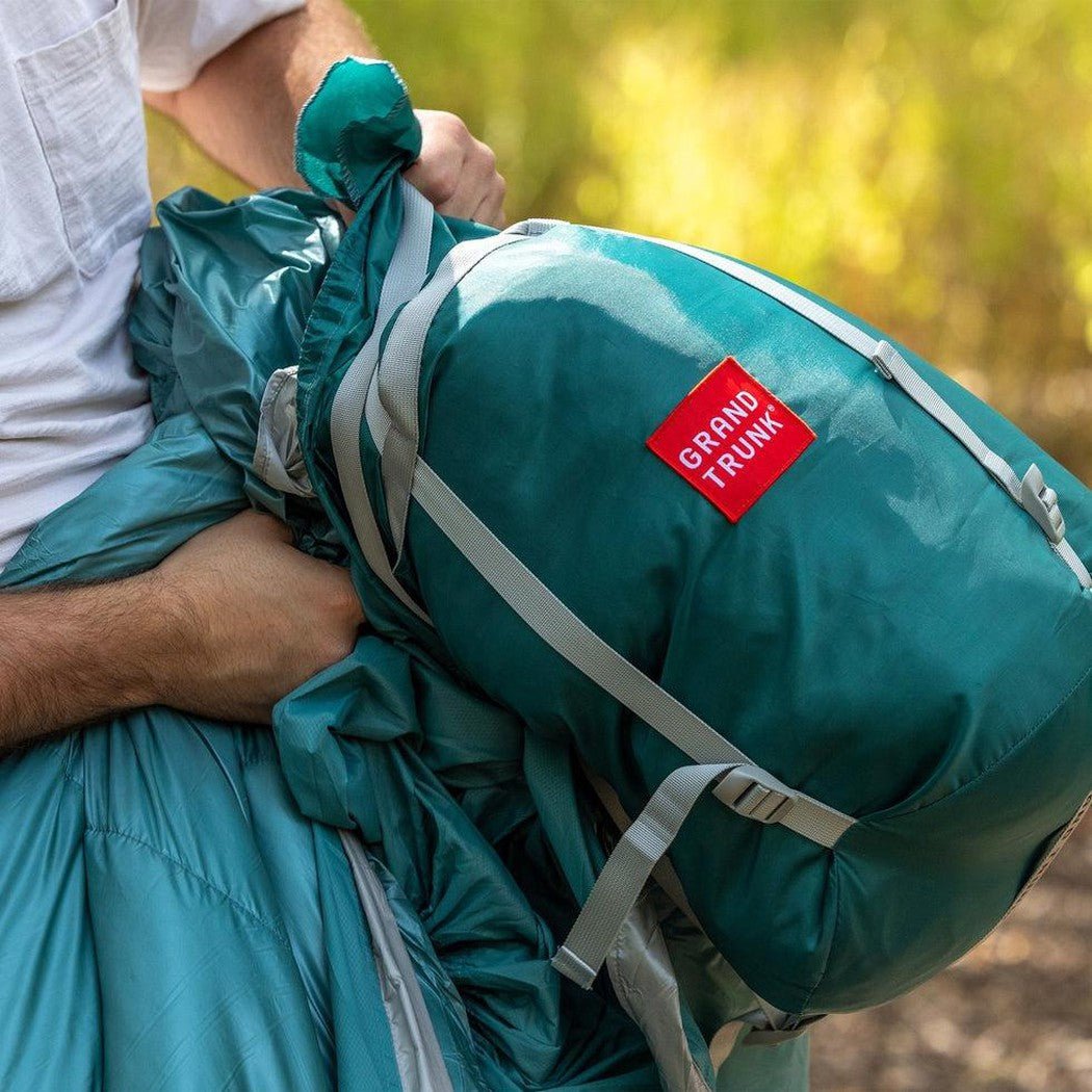 A person holds a teal Grand Trunk Evolution Synthetic Hammock with a red logo, against a blurred green background.