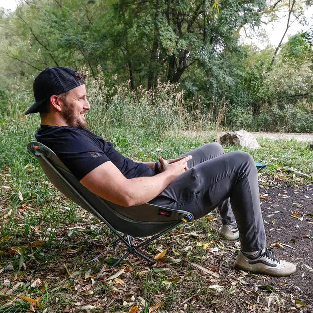 A man in a black Grand Trunk Mantis chair sits outdoors on a leafy ground with trees in the background.