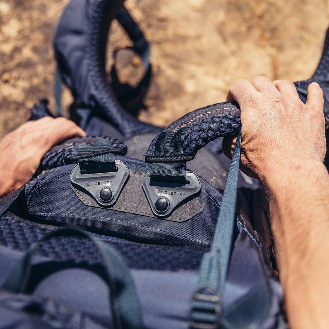 A person’s hands grip the handle of the Gregory Baltoro 65 backpack, which has two metal buckles and a blue strap, against a brown outdoor background.