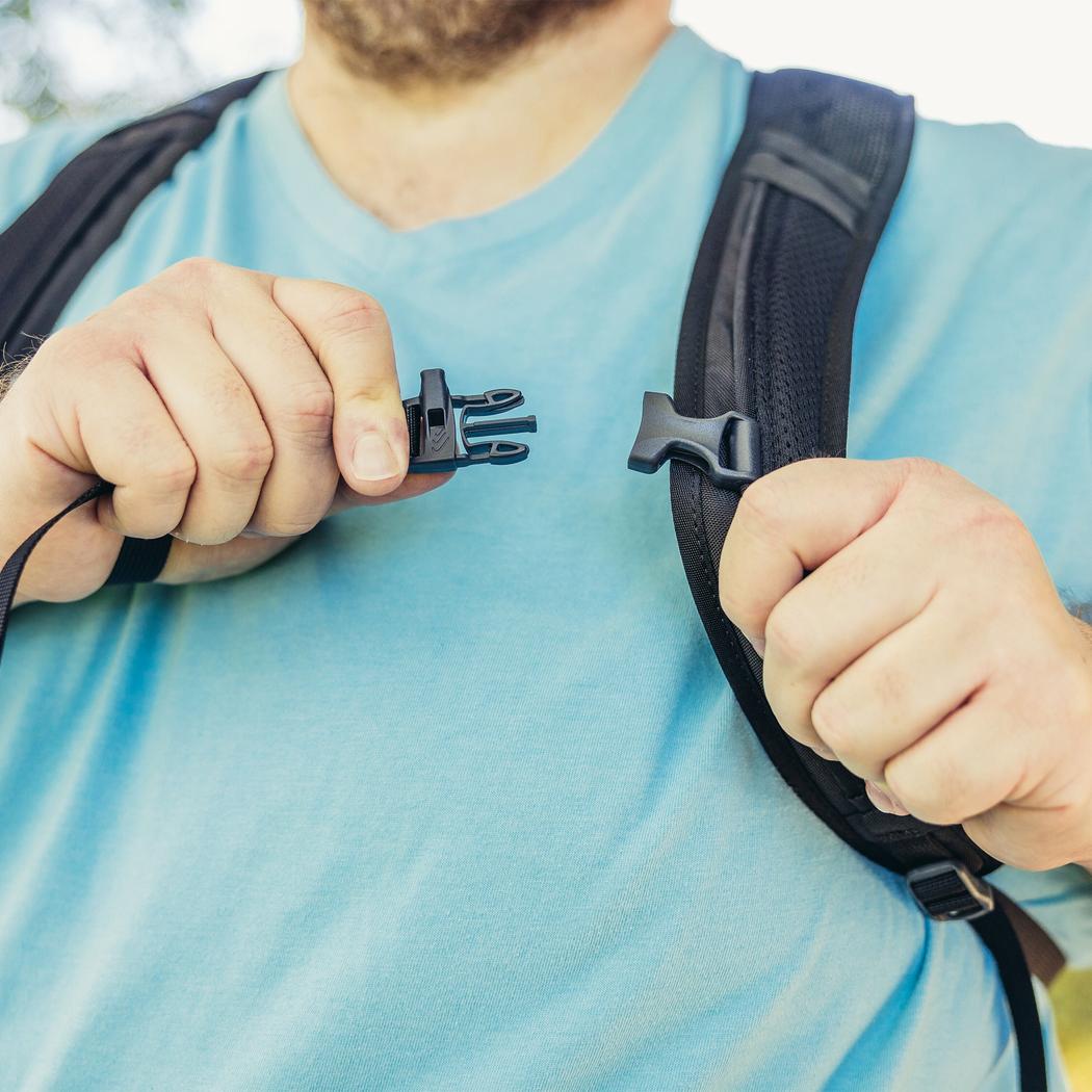 A man in a light blue shirt adjusts the buckle of a Gregory Nano 16 Plus backpack with a charcoal/gray shoulder strap.