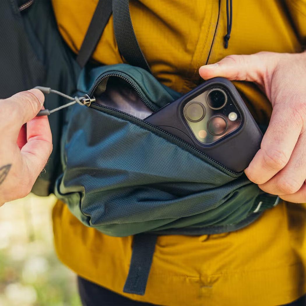A person in a yellow jacket holds a phone in a Cascade Green Gregory Paragon 70 backpack with a zippered pocket.