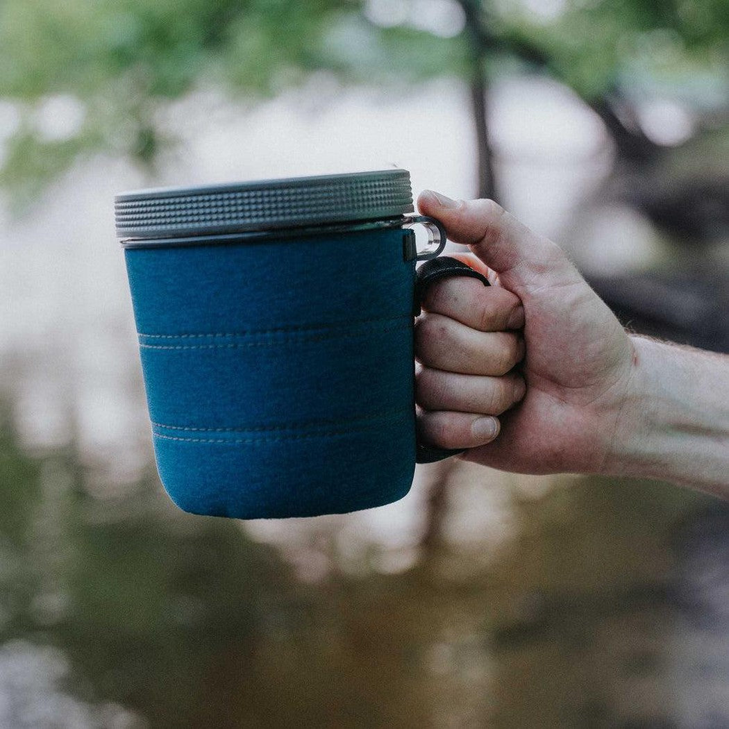 A hand holds the GSI Outdoors Infinity Fairshare Mug in Green, showing the neoprene sleeve and metal lid against a blurred outdoor background.