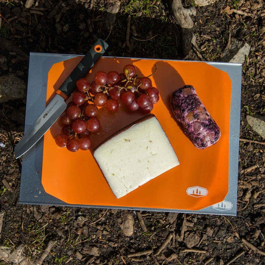 A GSI Outdoors Rollup Cutting Board Knife Set on an orange cutting board with grapes, cheese, and a knife, placed on a gray surface outdoors.