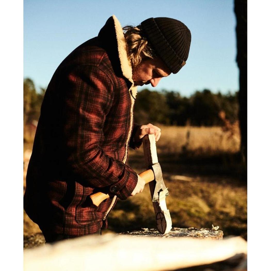 A man in a plaid jacket and beanie uses the Helko Werk Rheinlander Hatchet to chop wood in a sunny outdoor setting.