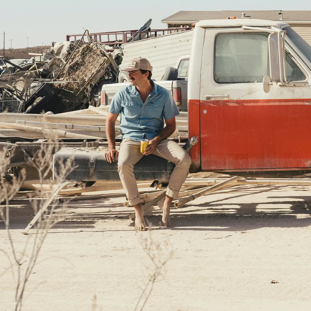 A man sits barefoot on a truck bed wearing Faded Khaki Howler Brothers Frontside 5-Pocket Pants in a desert junkyard setting.