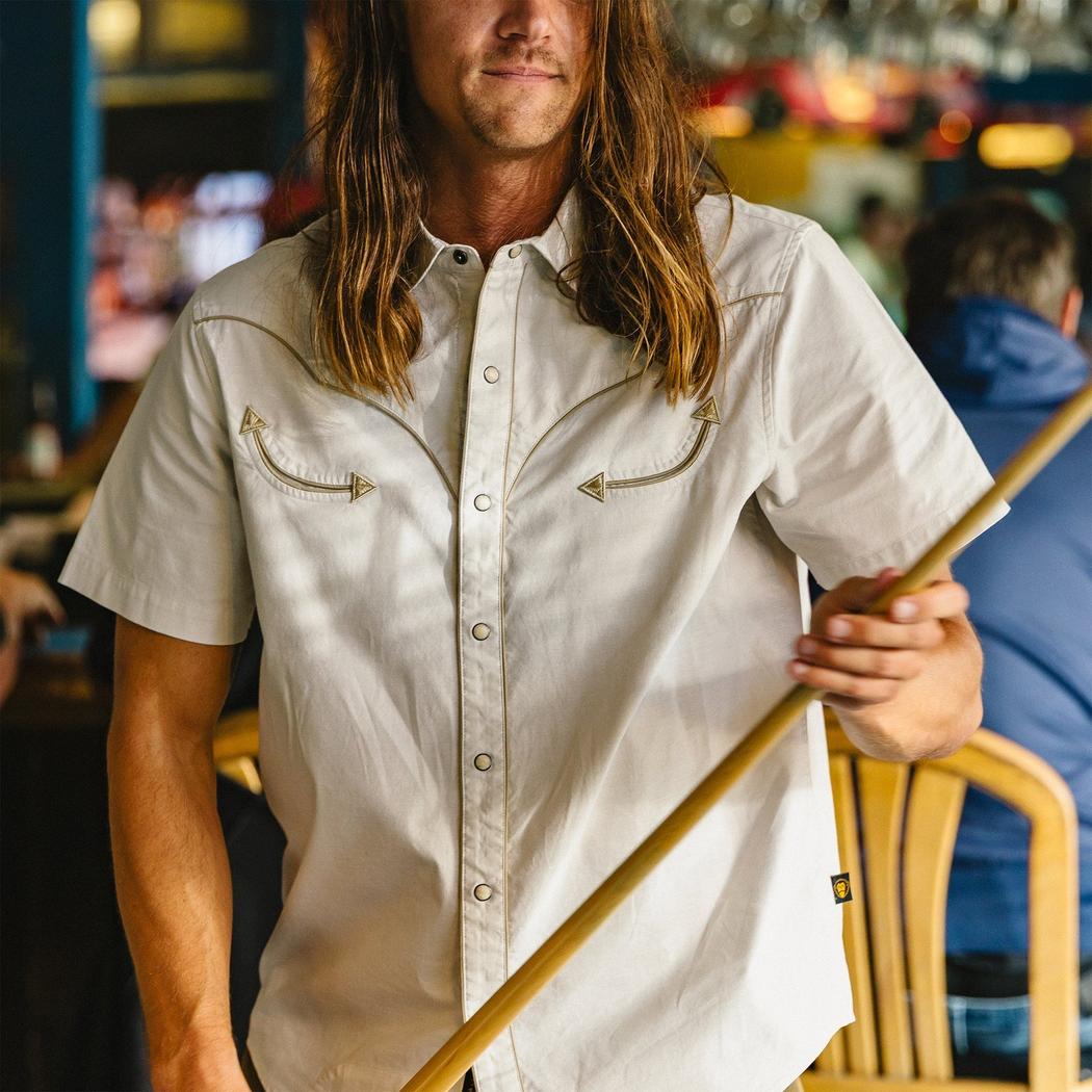 A man with long hair wears the Howler Brothers Men's Broncbuster Snapshirt in Sand, holding a pool cue in a bar setting.