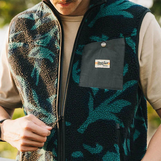 A man wears the Howler Brothers Men's Chisos Fleece Vest in Forgotten Forest : Antique Black, showing the front pocket and zipper.