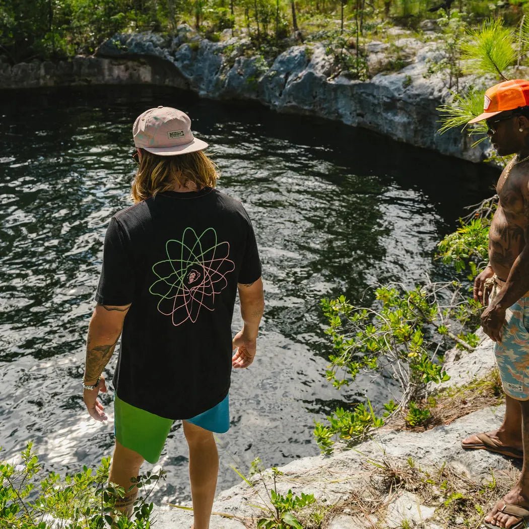 Two men stand by a rocky pool, one wearing the Howler Brothers Men's Cotton Pocket T in Black with a colorful atom graphic on the back.
