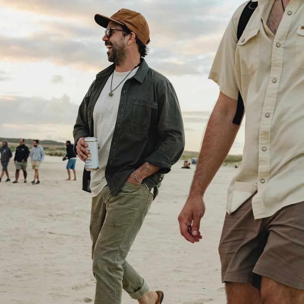 A man wearing the Anthracite Howler Brothers Mens HB Trade Shirt Long Sleeve walks on a beach at sunset with other people in the background.