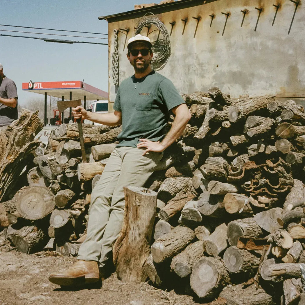 A man in Bone White Howler Brothers Men's Spectrum Pocket T leans on a log with a stack of firewood behind him.