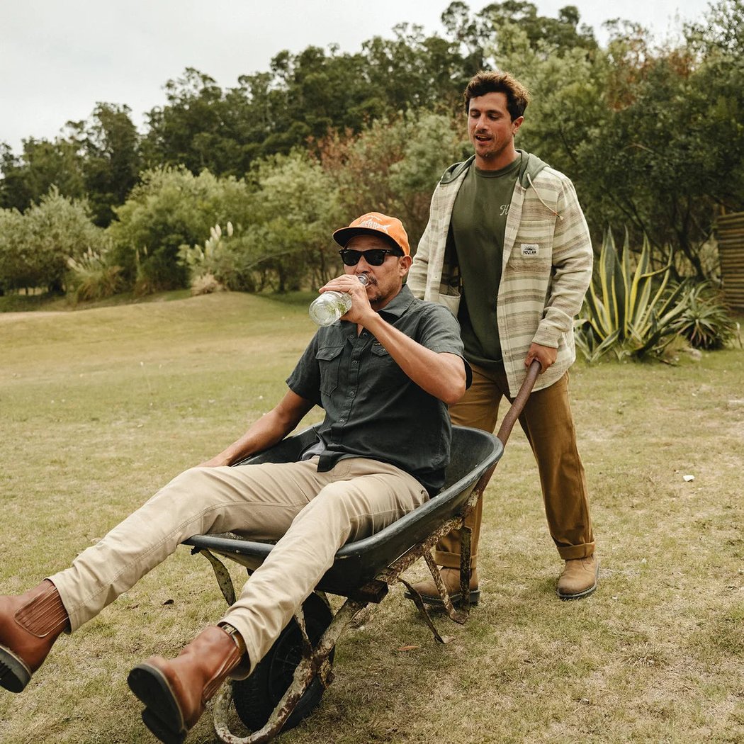 A man sits in a wheelbarrow wearing the Howler Brothers Salvo Hooded Blanket Flannel while another pushes it in a grassy outdoor setting.