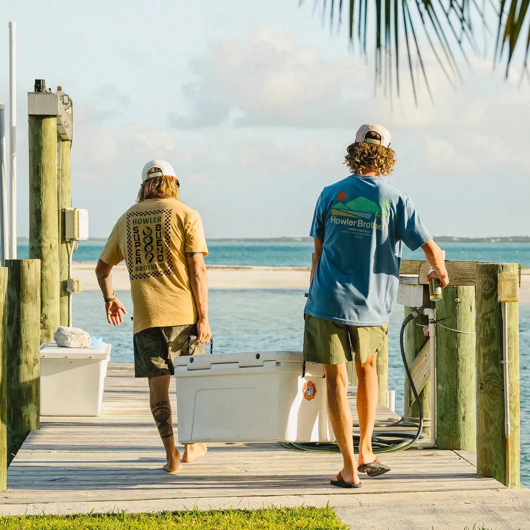 Two men walk toward the water wearing Howler Brothers Select T shirts, one in Antique Black and one in blue, on a wooden dock.