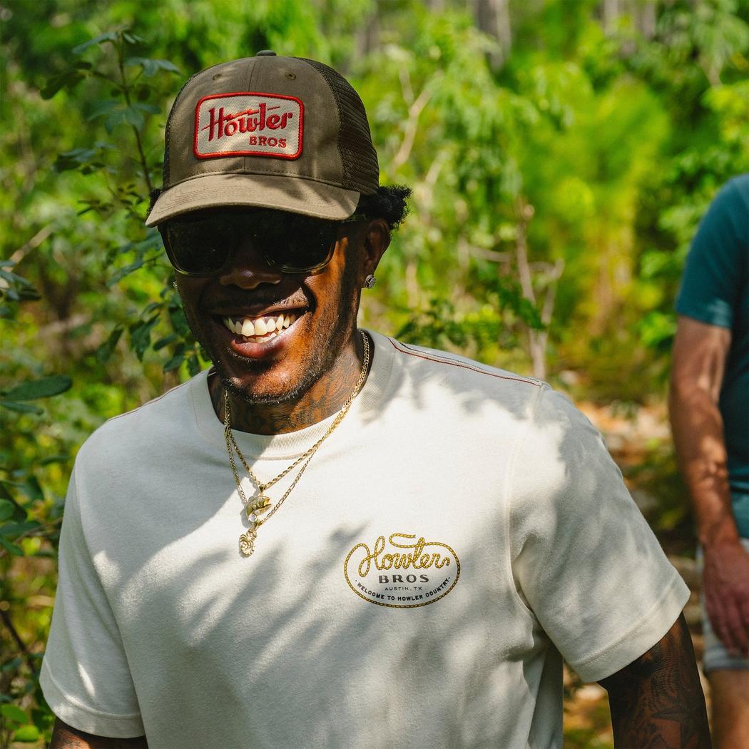 A smiling model wears a Howler Brothers Standard Hat in Howler Electric Fatigue color, with a forest background.