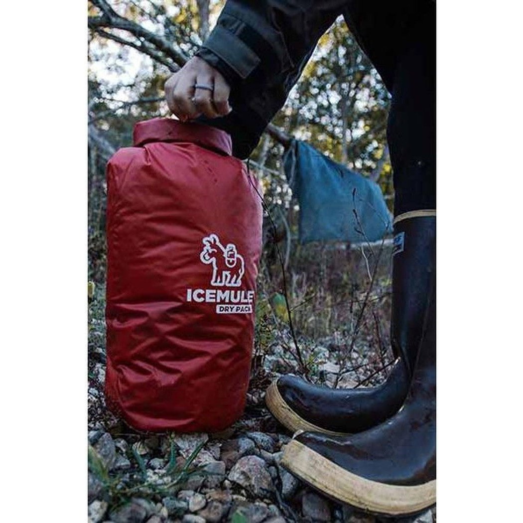 A person in black rain boots holds a red IceMule Dry Pack on rocky ground with trees in the background.