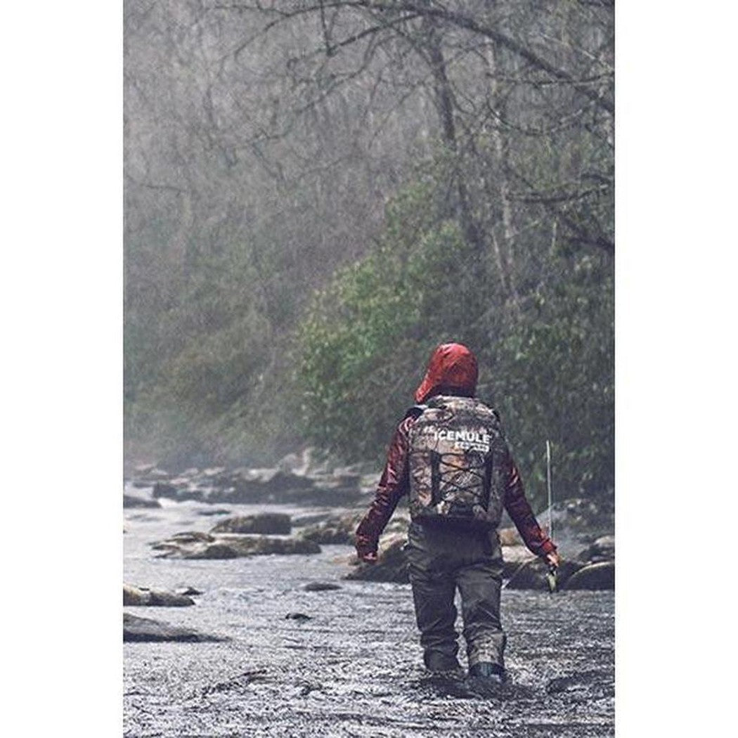 A person wearing a red hood walks through a stream with a Black IceMule Coolers Pro Cooler Large 23L on their back, surrounded by trees and rocks.