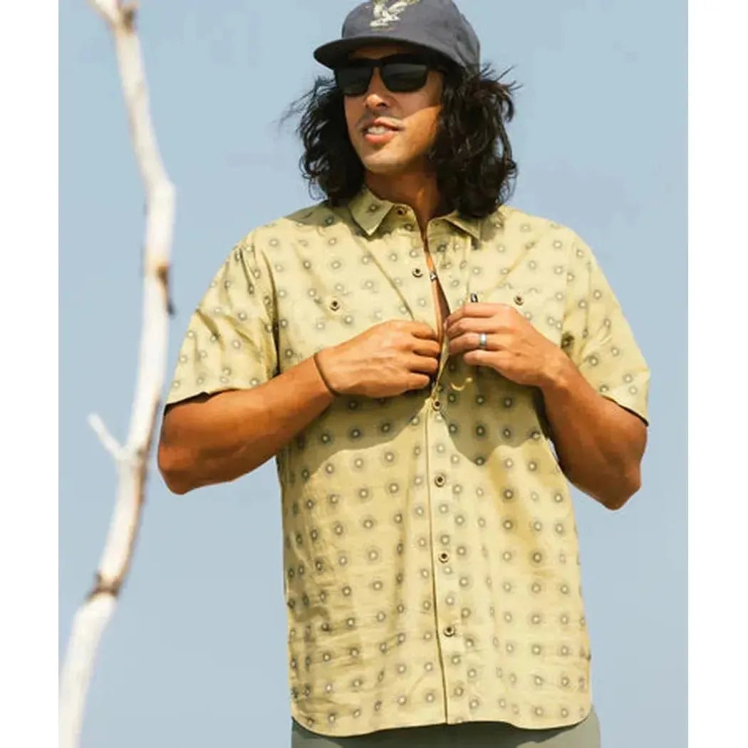 A model wears the Jetty Men's Seabrite Cotton Shirt in Navy, buttoning it against a light blue sky with a bare branch visible.